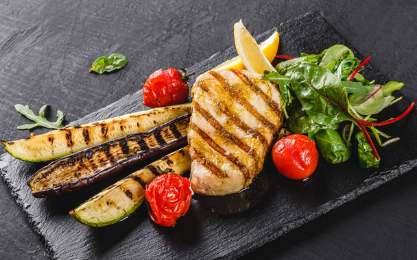Grilled fish steak garnished with salad of spinach and grilled vegetables on shale background over dark background. Hot fish dish. - Photo, Image