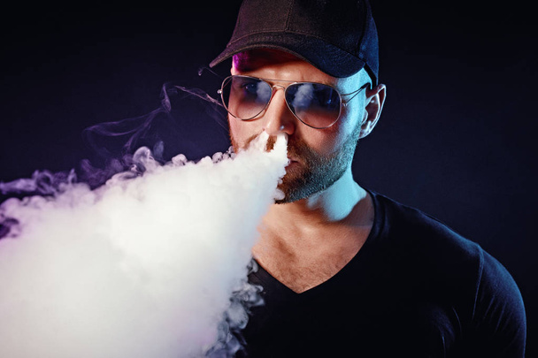 Men with beard in sunglasses vaping and releases a cloud of vapor - Фото, изображение