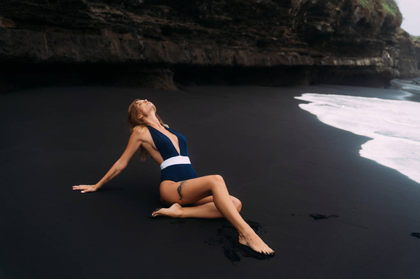 Sexy girl with big breasts in blue swimwear relaxing at beach with black sand. - Фото, изображение