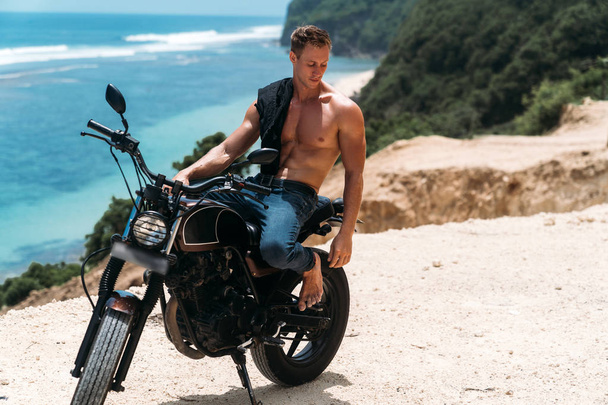 Sexy athletic man with perfect naked body sitting on motorbike, ocean waves and beautiful mountains on background - Foto, Imagem