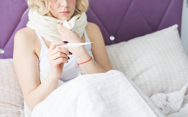 Beautiful blond woman sitting in bed with a thermometer and a scarf wrapped around the neck and a handkerchief. illness and health at home - Photo, Image