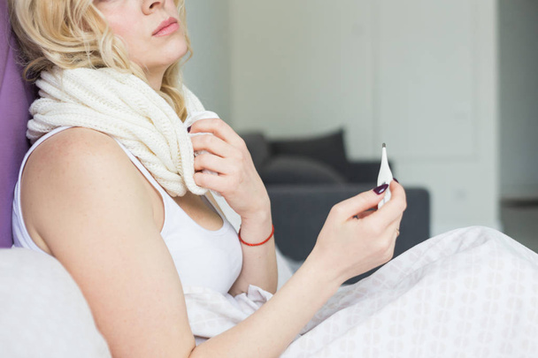 Красивая блондинка, сидящая в постели с термометром и шарфом, обернутым вокруг шеи и платком. болезнь и здоровье на дому
 - Фото, изображение