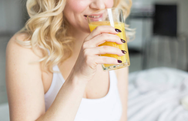 Attractive blonde woman sits on the edge of the bed at home and holds a stocker with orange juice in her hand. Morning mood and vigor - Φωτογραφία, εικόνα