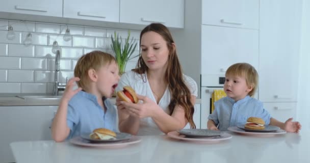 Une jeune belle mère en robe blanche avec deux enfants sourit et mange des hamburgers frais dans sa cuisine. Famille heureuse nourriture maison aliments sains
 - Séquence, vidéo