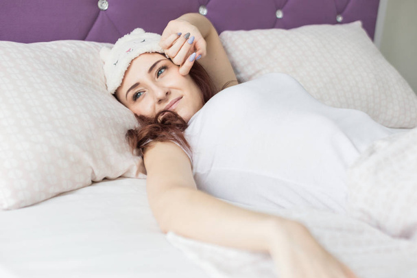 An attractive young brunette girl with brown hair wakes up and sips while yawning in her bed in a sleep mask - Photo, image