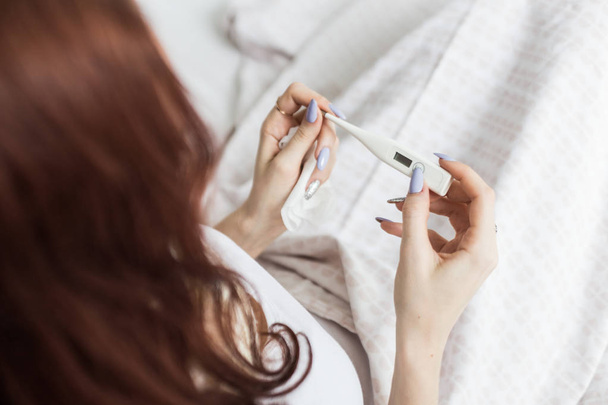 Sick woman with headache is sitting in bed. A sick girl with a seasonal infectious flu holds a thermometer in her hand and is covered in a blanket while lying in bed with a high fever. - Photo, Image