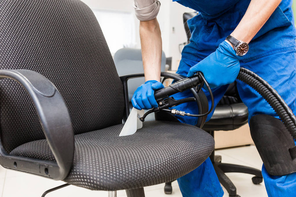 Young man in workwear and rubber gloves cleans the office chair with professional equipment. - Photo, Image
