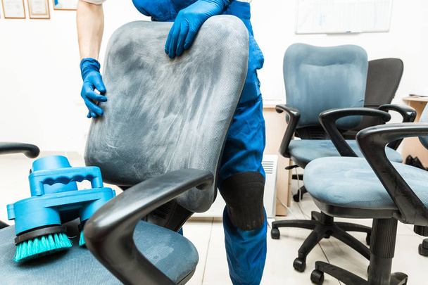 Young man in workwear and rubber gloves cleans the office chair with professional equipment. - Fotó, kép