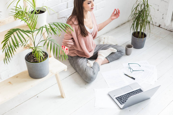 Young happy woman sits on the floor in a yoga pose in a bright apartment and works behind a laptop, a freelancer girl meditates while working. stress limit. - Фото, изображение