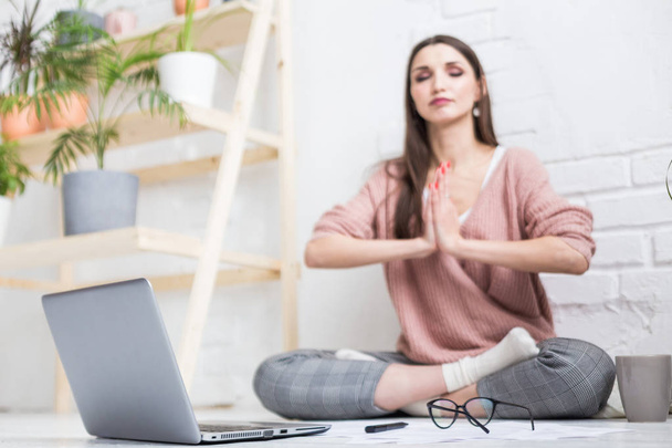 Eine junge glückliche Frau sitzt in einer hellen Wohnung in Yoga-Pose auf dem Boden und arbeitet hinter einem Laptop, ein freischaffendes Mädchen meditiert während der Arbeit. Spannung und Inspiration - Foto, Bild