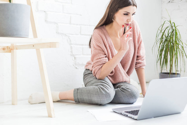 A young happy woman sits on the floor in a bright apartment or office interior and works at a laptop, freelancer girl at work, the concept of distance learning students, paperwork and online learning - Foto, Bild
