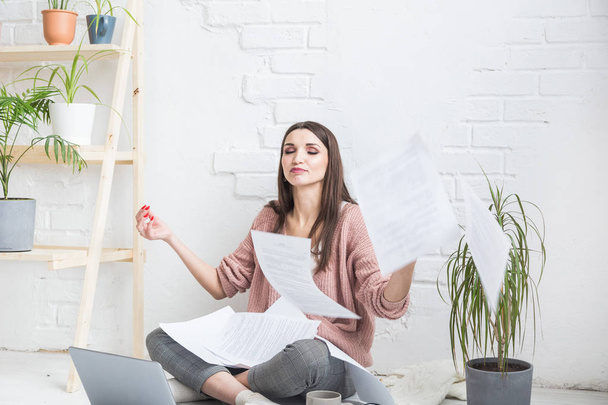 A young happy woman throws up a bunch of pieces of paper and documents sitting near the laptop, the freelance girl is happy and in a good mood. excitement and inspiration - Foto, Imagen