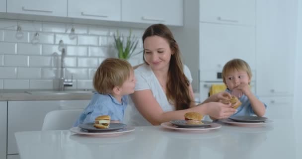 Una joven y hermosa madre en un vestido blanco con dos hijos sonríe y come hamburguesas frescas en su cocina. Familia feliz comida casera alimentos saludables
 - Metraje, vídeo