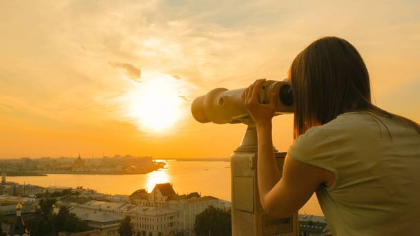 Νεαρή γυναίκα που κοιτάζει μέσα από το τουριστικό τηλεσκόπιο, εξερευνώντας το ιστορικό τμήμα της πόλης - Φωτογραφία, εικόνα