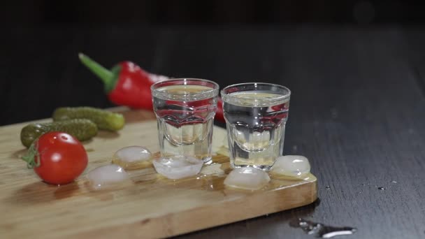 Vodka in shot glasses on rustic wood board. Adding ice cubes. - Imágenes, Vídeo