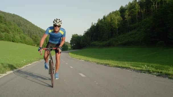 Slow Motion: atletische jonge mannelijke pedalen langs lege weg die door de natuur loopt. Wielrenner met zonnebril glimlacht als hij rijdt zijn fiets door het prachtige platteland. Professionele mannelijke Biker Racing. - Video