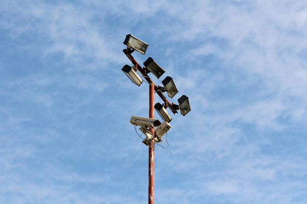 Ψηλό μεταλλικό πόλο με πολλαπλούς ανακλαστήρες φωτός και κάμερα ασφαλείας που συνδέονται με ηλεκτρικά καλώδια σε συννεφιασμένο γαλάζιο φόντο του ουρανού - Φωτογραφία, εικόνα