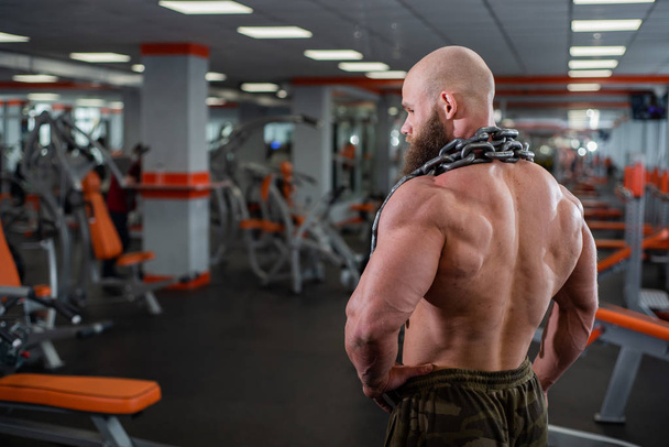 A bald, bearded bodybuilder with large relief muscles holds a heavy thick chain in his hands. Bright gym. - Photo, Image