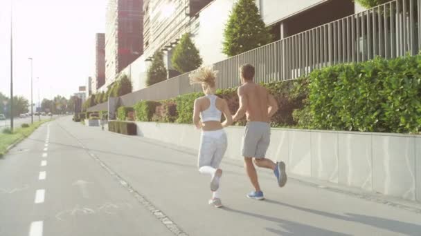 SLOW MOTION, LENS FLARE Couple bonds while jogging through city on a sunny spring afternoon. Caucasian couple runs along concrete pavement on a sunny day. Man and woman with athletic physiques running - Séquence, vidéo