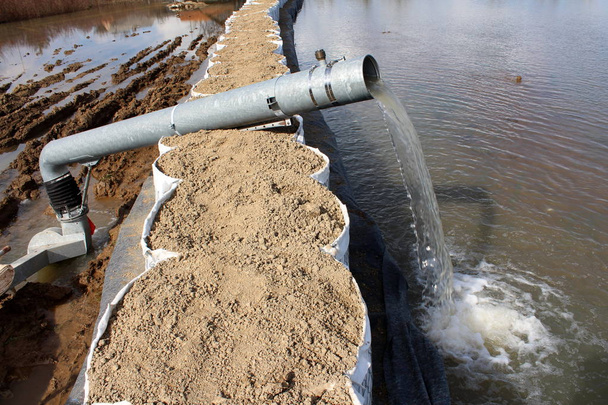 Bomba de água bombeando água de inundação sobre barreiras de caixa de areia altas através de mangueira de metal grande no dia ensolarado de inverno
 - Foto, Imagem