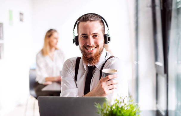 A portrait of young businessman with headphones and coffee in an office. - Foto, imagen