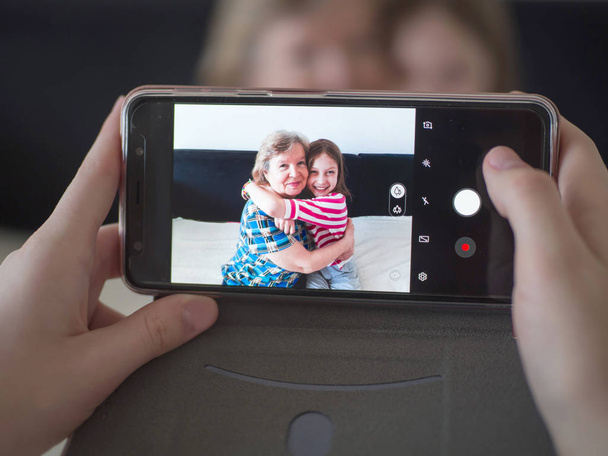 Joyful grandmother and granddaughter hugging at home, making photos on the phone - Photo, image