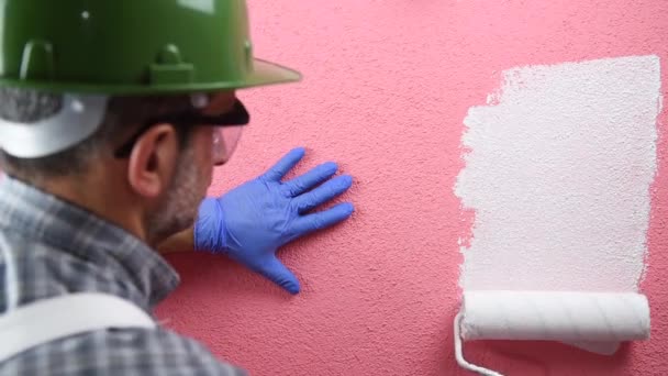 Kaukasische huisschilder werknemer in witte overalls, met helm en een beschermende bril. Met de roller schildert de roze muur met witte verf. Bouw. Beelden. - Video