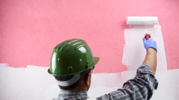 Caucasian house painter worker in white overalls, with helmet and protective goggles. With the roller paints the pink wall with white paint. Construction industry. Footage. - Footage, Video