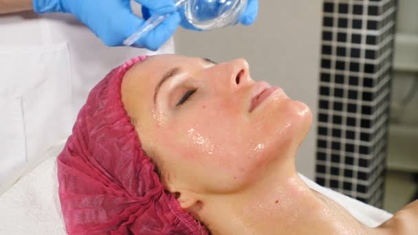 Esthéticienne féminine faisant le masque facial cosmétique au client féminin au centre de cosmétologie. Fermez-vous vers le haut de la fille assez caucasienne obtenant le traitement cosmétique sur son visage. rapprocher. Hd - Séquence, vidéo