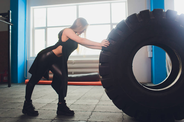 Fit female athlete working out with a huge tire, turning and carry in the gym. woman exercising with big tire. - Фото, изображение