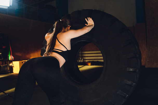 Fitness woman flipping wheel tire in gym. Fit female athlete working out with a huge tire. Back view. Sportswoman doing an strength exercise training. - Foto, imagen