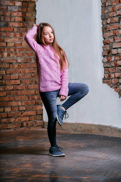 Portrait little fashion kid girl on brick wall background. Model tests young caucasian teen girl in pink sweater posing at studio loft,urban,street style. Teenager raising hand up holding hair - Foto, Imagen