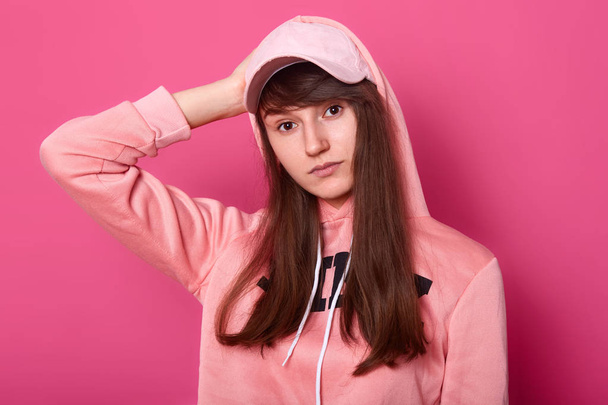 Image of brunette girl looks at camera with serious and thoufgtfull facial expression, wearing casual pink hoody and cap isolated on rose background, teenager poses in photo studio. Modeling concept. - Foto, afbeelding