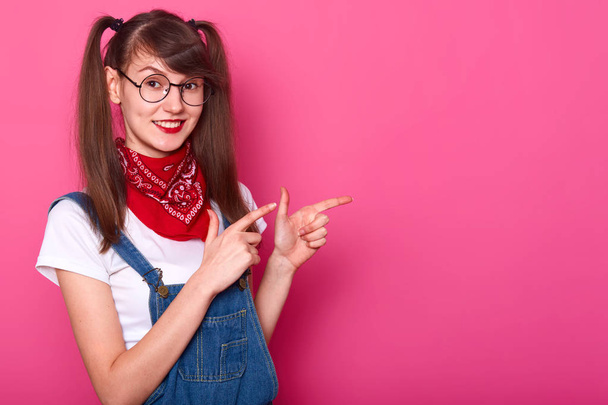 Smiling slim girl makes gesture with forefingers, showing the other way, looks pleased. Happy joyful young girl pigtails wears denim overalls, white t shirt, red bandana, fashionable spectacles. - Фото, изображение