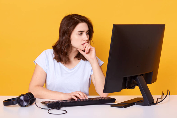 Studio shot of pleasant looking student with concentrated look, wears casual outfit, poses against yellow background. Beautiful woman working hard, earns money online, keeps hand under chin. - Foto, Bild