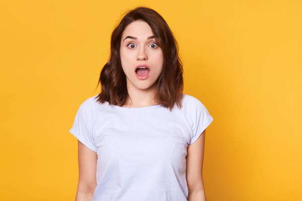Astonished slender young lady being impressed by latest news, stands with widely open mouth and brown eyes, looks surprised, has very emotional facial expression. Copy space for advertisement. - Foto, imagen