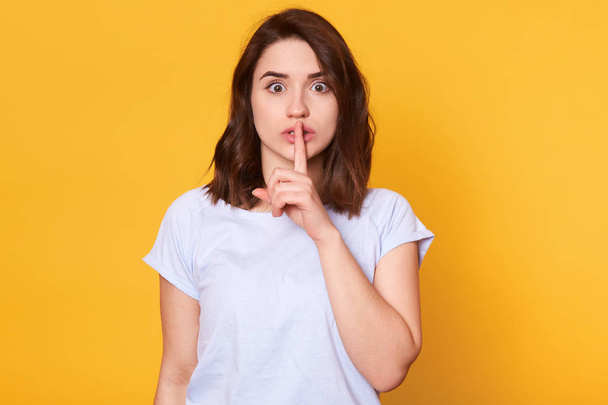 Slim attractive young lady stands straight isolated over bright yellow background, holding forefinger finger on her lips, making hint of being silent, opens her eyes wide, wears white casual t shirt. - Фото, зображення