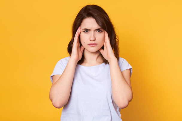 Woman with headache, has problem, lady standing with pain, keeps both hands on temples. Brunette female half length portrait. Isolated over yellow background. Human emotions, facial expression concept - 写真・画像