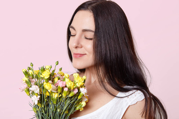 Close up portrait of black haired adorable young lady standing with closed eyes, holding early spring flowers, enjoys fantastic smell, looks delighted and peaceful. People and emotions concept. - Fotoğraf, Görsel