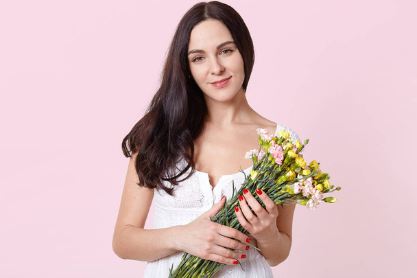 Portrait of smiling sincere young model standing isolated over light pink background, holding colourful spring flowers in hands, wearing white dress, looks satisfied. People and emotions concept. - Photo, Image