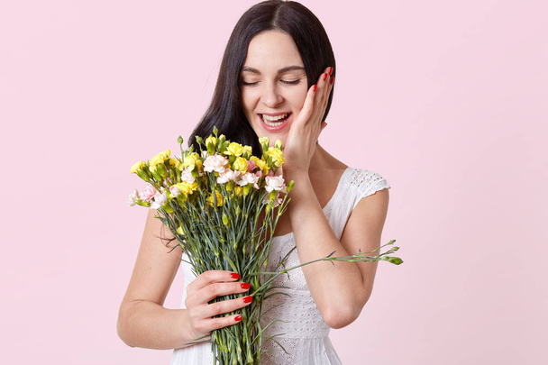 Laughing attractive young brunette model with closed eyes holding beautiful flowers in one hand, touching her face with other one, enjoys spending leisure time. Copy space for advertisement. - Valokuva, kuva