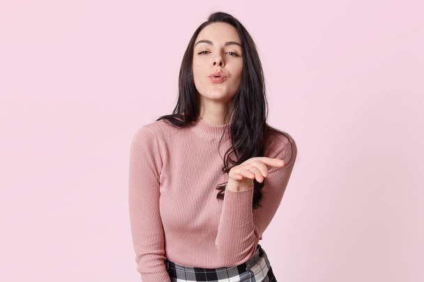 Portrait of tender cute romantic brunette girl making gesture, having pleasant facial expression, blowing kiss, posing over light pink background in studio. People, youth and emotions concept. - Fotografie, Obrázek