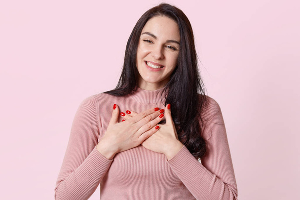 Adorable attractive young woman crosses hands on her chest, getting compliments, looks moving, wearing light pink jumper. People, communication and beauty concept. Copy space for advertisement. - Foto, Imagen