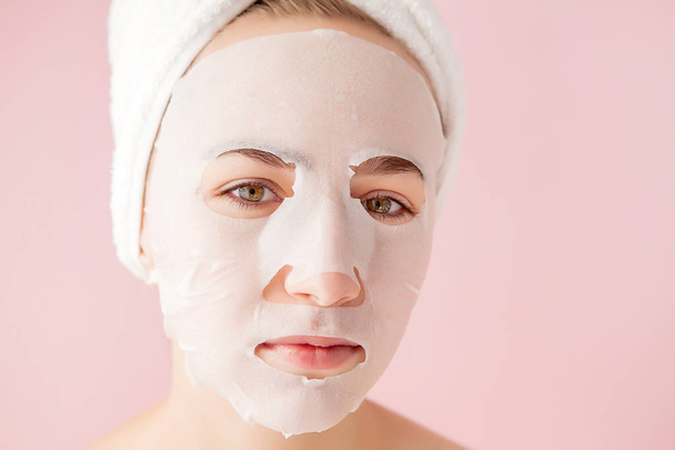 Beautiful young woman is applying a cosmetic tissue mask on a face on a pink background. Healthcare and beauty treatment and technology concept - Foto, imagen