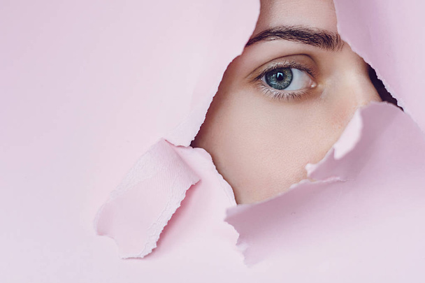 close-up of a female eye that looking is seen through a hole in a pink paper. Professional cosmetics and facial care. Bright colored background and a gap slot in the paper - Фото, изображение