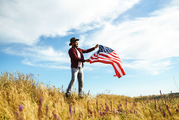 4 lipca. Czwarty lipca. Amerykanin z flagą narodową. Flaga Amerykańska. Dzień Niepodległości. Święto patriotyczne. Mężczyzna ma na sobie kapelusz, plecak, koszulę i dżinsy. Piękne światło zachodu słońca.  - Zdjęcie, obraz