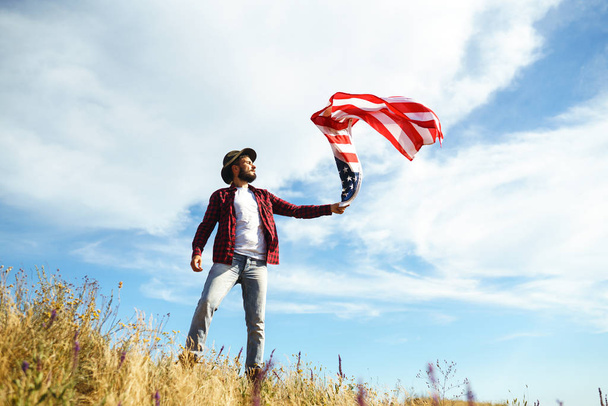 4th of July. Fourth of July. American with the national flag. American Flag. Independence Day. Patriotic holiday. The man is wearing a hat, a backpack, a shirt and jeans. Beautiful sunset light.  - Foto, imagen