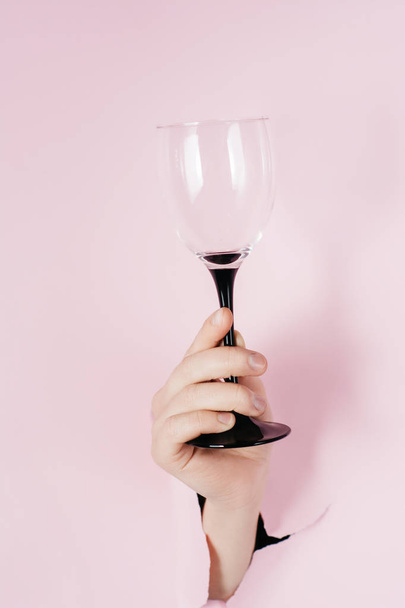 Creative images of female hand holds wine glass is seen through a hole in pink paper. Bright colored background and a gap slot in the paper. Holiday and birthday concept. - 写真・画像