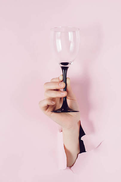 Female hand holds wine glass is seen through a hole in torn pink paper. Overhead view, copy space. Holiday and birthday concept. - Foto, imagen