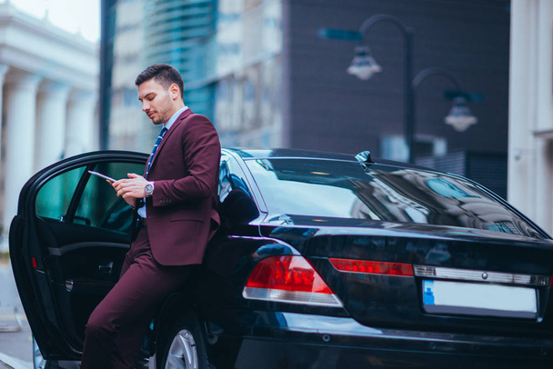 Knappe jonge zakenman leunend op zijn limo en glimlachend tijdje - Foto, afbeelding